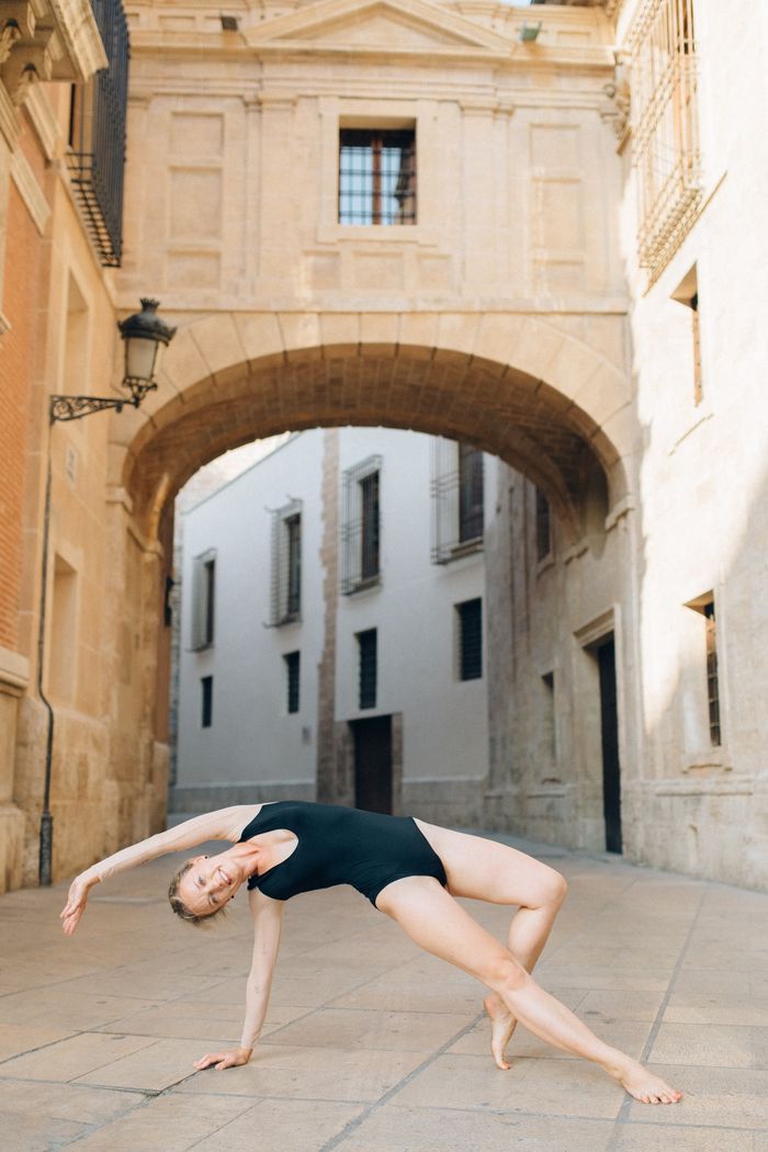 A woman in a black bodysuit executing a yoga pose on an urban street with historical architecture.