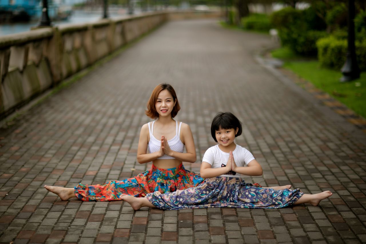 A mother and daughter performing yoga on a paved path, showcasing togetherness and wellness.