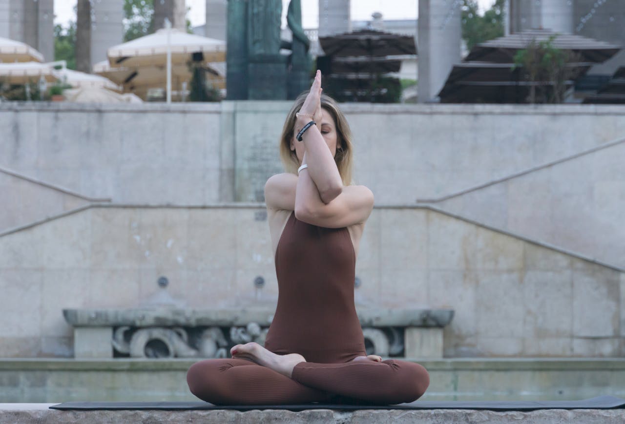 A young woman meditates in a yoga pose by an outdoor fountain, promoting relaxation.